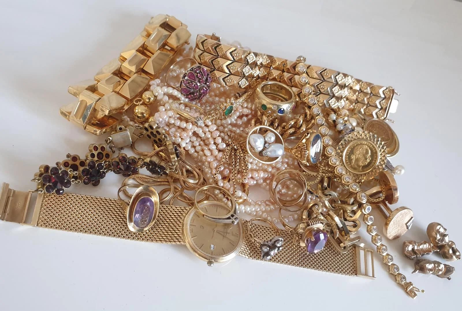 Goldene Armbänder, Ringe mit Farbsteinen, Perlenketten, Goldmünzen und eine goldene Armbanduhr auf hellem Grund
